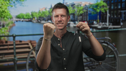Man clenches fist and holds car keys on a street in amsterdam by a canal bridge; triumph...
