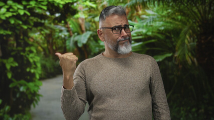 Man pointing thumb over shoulder at a green forest path, wearing glasses and grey beard with knit...