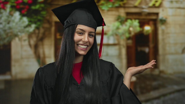 Hispanic woman in graduation cap smiling and pointing to her palm while standing outdoors against a charming rustic building background.