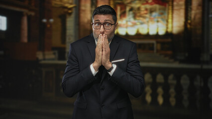 Man in suit with hands covering mouth and glasses standing in church building amid altar and pew,...