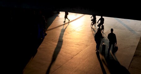 Dramatic silhouettes of people walking under a bridge with long stretched shadows at sunset. Strong golden light creates an atmospheric, cinematic mood evoking mystery, solitude, and contemplation.