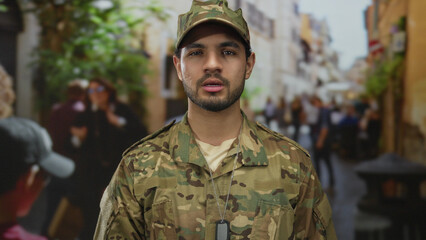 Man in camouflage military uniform stands upright and looks ahead with steady gaze on crowded street; service honor commitment duty.