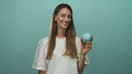 Woman smiling holds blue piggy bank in right hand wearing white t shirt in mint studio; savings...
