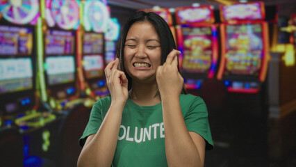 Chinese woman in a volunteer t-shirt crosses fingers optimistically inside a vibrant casino with slot machines in the background displaying bright lights and colors.