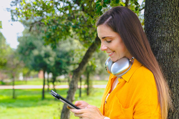 Happy woman wearing yellow shirt leaning on tree using smartphone and smiling outdoors in sunny green park, enjoying communication, technology and nature lifestyle concept with positive expression