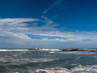 Dynamic Atlantic Ocean waves crashing against rugged rocky formations under a dramatic blue sky with white clouds in Essaouira, Morocco