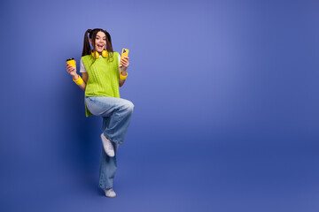 Young woman in bright green vest with headphones holds phone and coffee against blue background for...