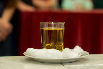 A clear glass filled with a yellow herbal drink rests on a white plate with cotton. People in the background chat and enjoy their time during a social gathering.