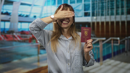 Young woman holding passport in indoor pool building, hand visible over passport, wearing striped...