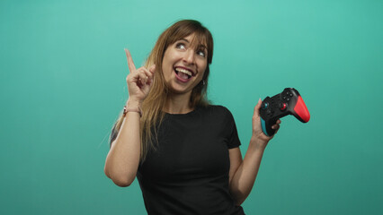 Woman holding colorful game controller points finger upward in teal studio smiling broadly; playful gaming joy.