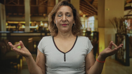 Woman making ok sign with raised hands while closing her eyes in a hotel building lobby wearing striped shirt; serenity.