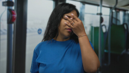 Woman in blue shirt with eyes closed covering face with hand inside a city bus cabin during morning commute while standing upright; fatigue.