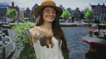 Fototapeta premium Hispanic teenager wearing summer hat smiles and points finger forward in street; joyful connection invitation.