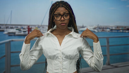 Woman points thumbs to chest at port boardwalk wearing white blouse and glasses, braids visible; confidence.