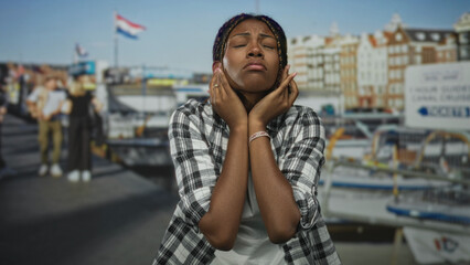 Woman covering ears with hands at building by the port and boats, wearing plaid shirt and wristband; frustration travel delay.