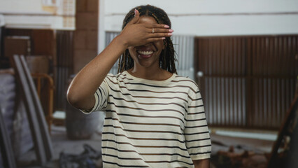 Woman smiling with hand covering eyes in a building resembling an indoor construction site, striped...