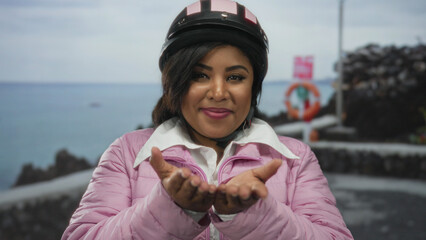 Young woman wearing helmet and pink jacket extends hands towards camera by the seaside promenade