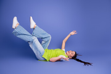 Young woman in bright lime knit top lies on blue studio backdrop with legs up and playful arms in a...