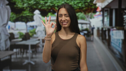 Fototapeta premium Woman making ok sign with right hand and winking on street terrace of a restaurant with wristwatch visible; approval casual.