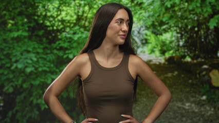 Fototapeta premium Woman with hands on hips in a green forest clearing smiling gently at viewer, bare shoulders and long hair visible; serenity confidence.