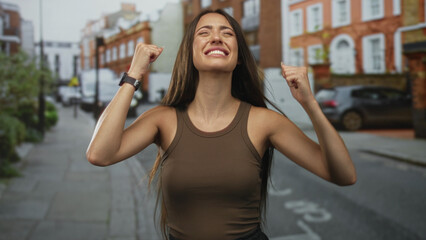 Young hispanic woman smiling and raising fists with bare arms on a city street in sleeveless top; personal success triumph.