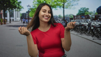 Fototapeta premium Young hispanic woman smiling and beckoning with both hands on a busy street beside parked bicycles wearing a red top and jeans; playful invitation.
