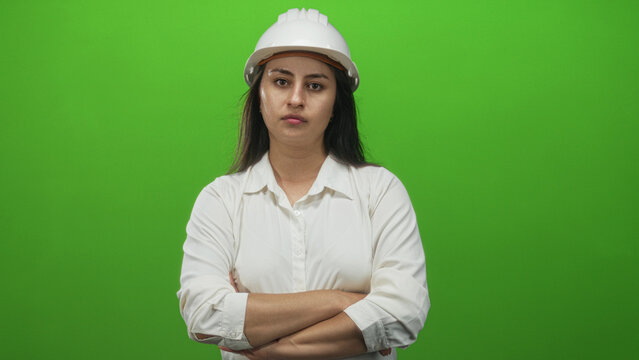 Young hispanic woman architect wearing white shirt and hardhat pointing with hand toward right in green studio; confidence.
