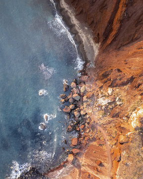 Aerial view of the stark, red cliffs plunging dramatically into the turquoise sea, the rugged coastline creating a mesmerising contrast of color and texture, Fira, Thira, Greece.