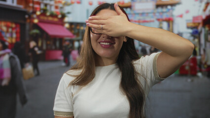 Woman covers eyes with hand in a colorful outdoor city street market bustling with lanterns and pedestrians; playfulness.