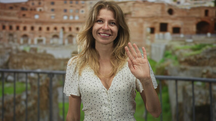 Fototapeta premium Woman smiling and waving right hand at roman ruins building with railing in foreground, casual dress and sunlight; greeting warmth.