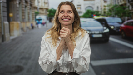 Fototapeta premium Woman with hands clasped at chest on a busy city street, smiling with folded fingers, wearing white shirt and belt in front of cars and building facades; hope.