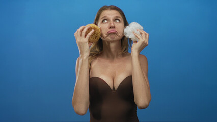 Woman holds natural sponge and white loofah to face showing deep cleavage in blue studio; pensive skincare.