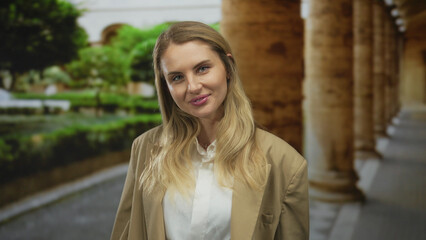 Fototapeta premium Woman smiling outdoors in a lush garden setting with arched architectural elements around, wearing a beige blazer and exuding confidence and warmth.