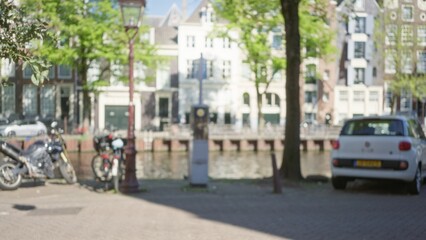 Blurred view of amsterdam canal with parked cars and bicycles under sunny skies, showcasing defocused architecture and greenery.