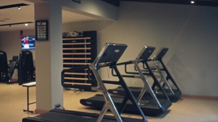 Blurred image of an indoor gym with multiple treadmills lined up beside a wall under soft lighting,...