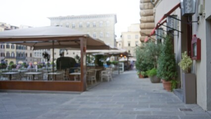 Fototapeta premium Blurred outdoor cafe plaza scene with soft bokeh, shallow focus on empty tables and potted plants, distant architecture out of focus; background backdrop copyspace calm.
