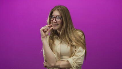 Young blonde woman wearing glasses pinches fingertips to show tiny measure in purple studio; playfulness.
