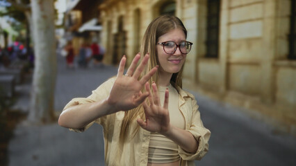 Fototapeta premium Blonde woman wearing glasses holds palm outward in stop gesture on urban street in front of building facade; discomfort.