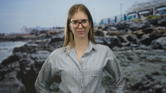 Blonde woman wearing glasses and striped shirt with hand on cheek by rocky seaside beach; doubt concern thought.