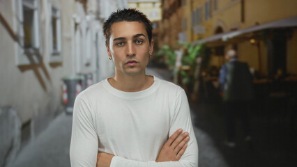 Young hispanic man in white shirt with arms crossed at a street terrace restaurant outdoors in...