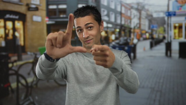 Young hispanic man making loser gesture with hand and pointing at camera in busy urban street; mockery.