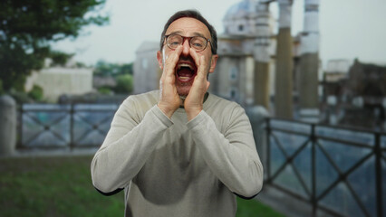 Man shouting energetically outdoors in urban setting with architecture background, wearing glasses and sweater, mid-shout against city landscape.