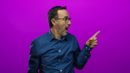 Mature man with glasses enthusiastically pointing on a vibrant pink background wearing a blue shirt.