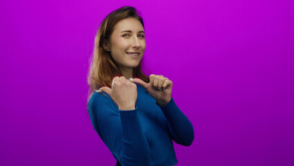 Young woman with long hair wearing a blue sweater pointing thumbs behind her while smiling and winking on an isolated vibrant pink background.