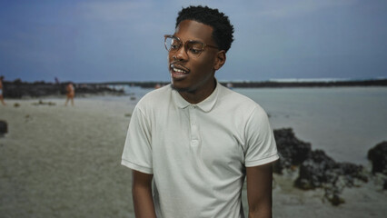 Young african american man wearing polo shirt and smiling with glasses on sandy beach near rock formation; serenity and new beginnings.