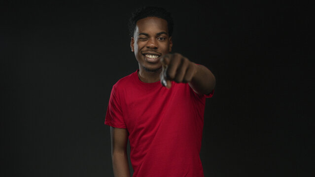 Friendly smiling young african american man pointing finger in studio against black wall wearing red shirt; confidence.