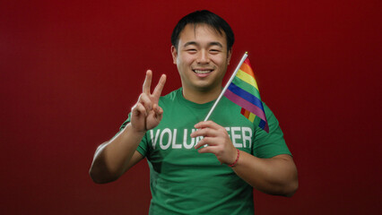 Young man holding rainbow flag and making peace sign in front of red wall showing pride and support...