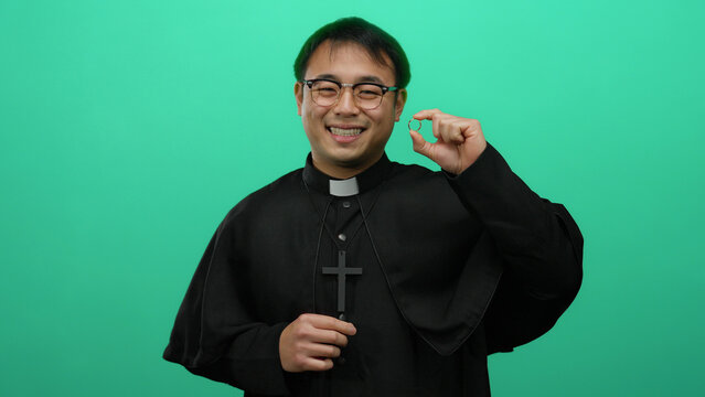 Priest smiling while holding ring on green background, suggesting proposal or marriage theme highlighting cultural aspect with asian man in focus.