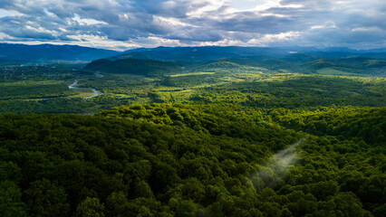 Drone shot over vast green valley with sunlight beams breaking through dramatic clouds, revealing forest textures, hills, and the beauty of pristine mountain wilderness