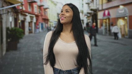 Fototapeta premium Young latin woman smiling while looking upward with head tilted on sunlit city street under soft natural light; optimism.